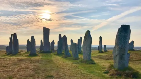 CHARLIE MCGINN A general view of the Callanish Stones. There are a number of grey standing stones of varying sizes. All of the stones are grey, but those in the background of the image look darker due to the shadows of the sun. The sun is above the tallest stone and is partially obscured by cloud. The sky is blue. To the right of the image, two clouds have crossed over in the sky in a Saltire pattern.