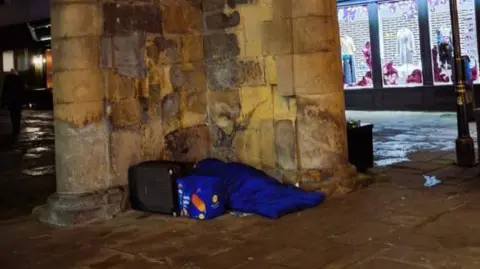 Shropshire Council A person in a blue sleeping bag in Shrewsbury. They are against an old wall and there is a shop lit up in the background. 