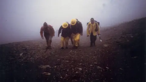David Needham Two firefighters climbing up a mountain in 1986 in full kit, with two people next to them