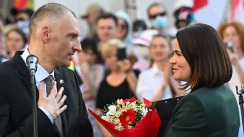 EPA Sergei Tikhanovsky and his wife, Belarusian opposition leader Svetlana Tikhanovskaya, look at each other smiling. She is holding a bouquet of red and white flowers and he is holding his hand to his heart.