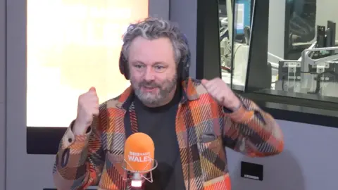 Michael Sheen who has grey hair and a grey beard, sat in a radio studio behind an orange microphone with black headphones on his head. He is wearing an orange and black chequered jacket