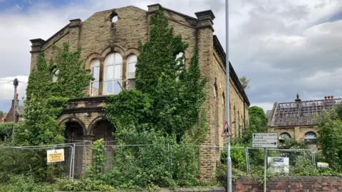 LDRS An old, run-down looking building with large circular windows on the side. There is greenery covering part of the exterior and a metal fence bordering some of the building. Next to the larger building is a smaller stone building which has part of its roof missing. 