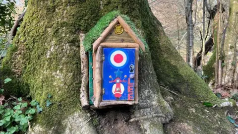 A small door painted blue and decorated with a red circle surrounded by white and the name Lee is attached to the base of a moss covered tree in woodland.