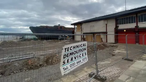 Construction site of gravel on flat ground next to a two-storey building with old vessel in the background