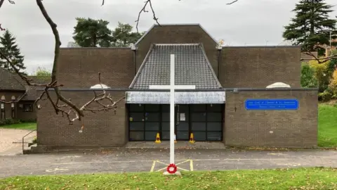 1970s stone-built church with glass entrance doors. A white cross stands in front of the entrance.