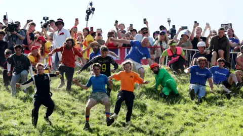 Getty Images A crowd of men launch themselves from the top of Cooper's Hill near Gloucester for the annual cheese rolling competition. It isa hot sunny day and the crowd at the top of the hill are filming on smart phones. 