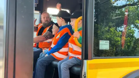 Contributed Three young boys wearing matching black caps, orange hi-vis jackets, blue jeans and different coloured jumpers sit inside the cab of a bin lorry. A man wearing a matching orange hi-vis jacket speaks to them from the driver's seat.