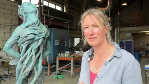 Artist Rebecca Hawkins, wearing a grey top, standing in her studio alongside the sculpture