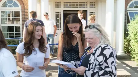 Peter Whittlesea/BBC Two students looking at their results with a teacher at the school. The two girls have long brown hair; one is wearing a white t-shirt, the other a polka dot black and white top. The teacher has whitish hair and is wearing a black and white linen shirt with black top underneath. 