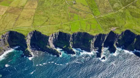 Getty Images An aerial view of the Cliffs of Moher in County Clare on the west coast of Ireland. Waves are gently breaking below the tall cliffs which stretch over a large distance. The ground behind the cliffs is fields crossed by several paths and trails.