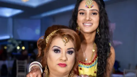 A woman with long dark hair, jewellery, and a yellow dress with her arm on the shoulder of a woman with ginger hair and jewellery