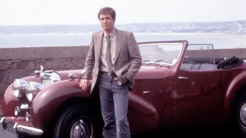 John Nettles pictured as Bergerac in the original series. He is seen learning against a dark red classic car while wearing a green tweed jacket with a beige shirt and green tie.