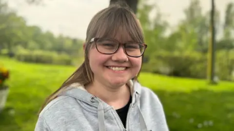 Hannah Fullerton from Buglife has glasses,  brown hair, tied back, and a fringe. She wears a grey hoody and is smiling widely. 
