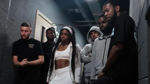 BBC/Naked TV/Andrew Efah Rap Game UK series six cast, left to right: CBliminal, Amani Steez, Layyah, Haydog, Only Zizou and Fumez the Engineer stand in a breeze-block hallway that's been painted black. 