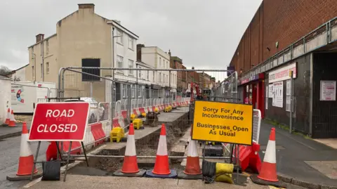 BBC Fences block the road, with signs saying 'road closed' and 'sorry for any inconvenience - businesses open as usual' either side. There are traffic cones at the front of the fence, and the road is flanked by properties.