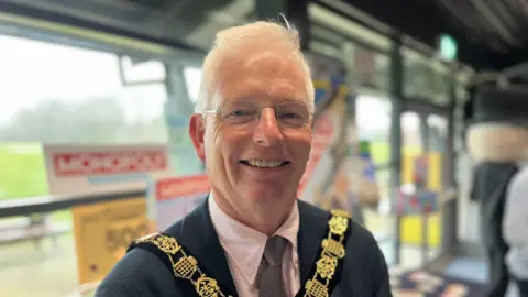 George Carden/BBC Mayor Sean McHale standing in front of the board game wearing his mayoral chain and a suit smiling at the camera