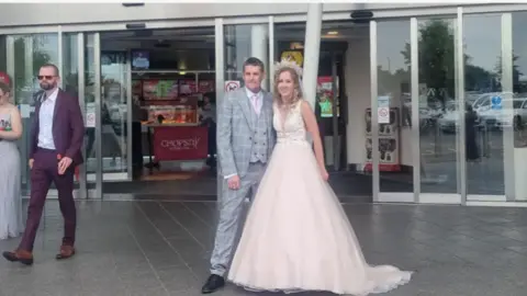 Stacey A groom and bride standing outside the service station. The bride is wearing a cream/ light pink wedding dress with a embroidery top. The groom is in a grey checked suit and a pink tie.  