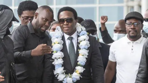 AFP Mozambique's main opposition leader Venancio Mondlane (centre) wearing a black suit and a flower necklace walks with people upon his arrival at the Maputo International Airport in Maputo