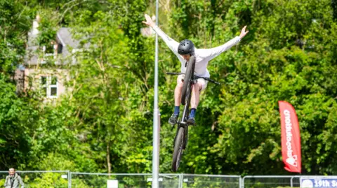 Pete Scullion A cyclist in white gear with a black helmet takes his hands off the handle bars of his bike while it is up in the air.