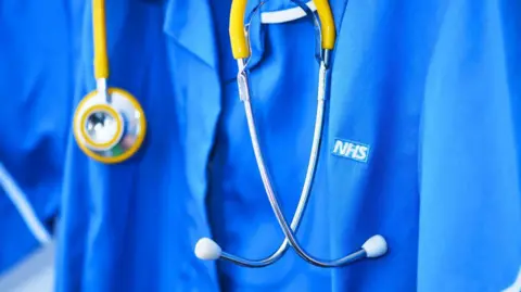 A close up of a healthcare working in blue scrubs with the white and blue NHS logo on the breast. There is a silver and yellow stethoscope around the person's neck