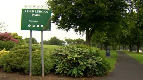 BBC A sign saying Lord Lurgan Park. There are bushes, and trees in view and a pathway.