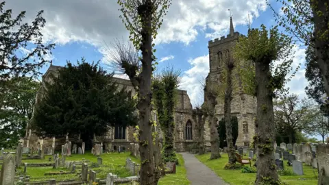 Nicola Taggart Elstow Abbey, showing a large stone building, with trees outside, and grave stones. There is a tower, and then a main building. 