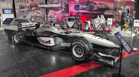Kate Bradbrook/BBC A black racing car with silver trim. The name David is seen in black writing on a white background on the side of the body, alongside "Computer Associates" and Mobil. There is an information stand in front of the car, which is parked on a black and red surface. There are display boards with pictures of racing cars behind it.