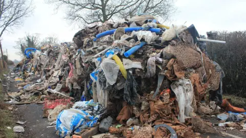 A huge pile of fly-tipped waste. The pile of waste visibly blocks the road.