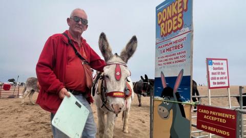 John Nuttall holding a set of scales next to a donkey on Skegness beach