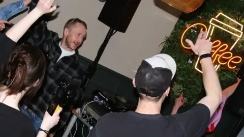 @scarlettamelie_photos People hold up in their arms in a coffee shop to show appreciation for the music Lauren is playing. One man is wearing a baseball cap. 