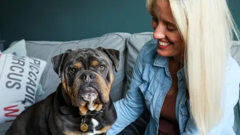 RSPCA A woman with straight blonde hair and a denim shirt sits on a sofa, smiling down at a bulldog perched to the left who looks at the camera. A cushion saying 'Piccadilly Circus' is behind him.