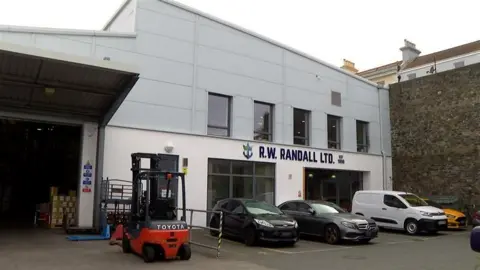 Exterior shot of the Randalls brewery offices. There are three cars and a van parked outside and a white van. A red Toyota forklift is also parked outside the building.