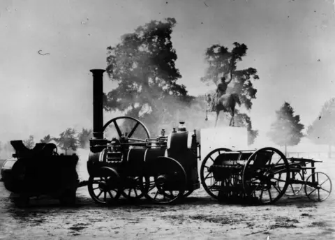 Hulton Archive via Getty Images A vintage black-and-white image of an early steam engine with a tall black chimney and large iron wheels. It is pictured with other agricultural machinery in what looks like a park, with trees and a statue of a horse and rider in the background.