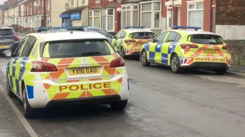 Three police cars are parked either side of a road, which has a row of terraced houses along one side.