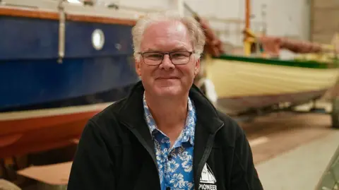Martin Giles/BBC Simon wears glasses, a black fleece and blue floral shirt underneath. He is sitting in front of a blue boat. He is looking directly at the camera and smiling. 