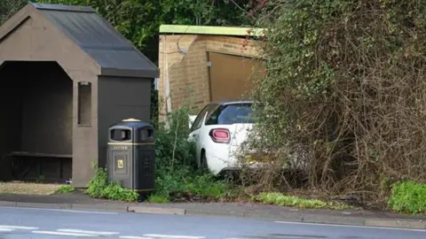 An image of the site in Pagham Road, near Nyetimber, where a car crashed into a gas substation. A white car can be seen crashed into the substation building, which is just beside a brown bus stop.