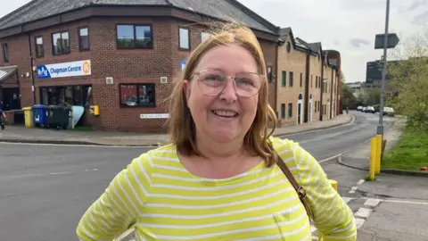Vikki Irwin/BBC Siobhan Cairns is wearing a yellow and white striped top and is standing on a street in central Ipswich. She is smiling at the camera and is wearing glasses. A two-storey brick building with a sign saying "Chapman Centre" is behind her.