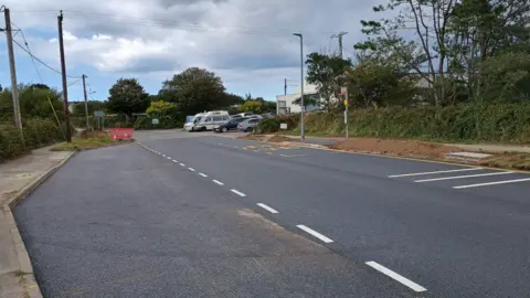 Newly resurfaced road with lane markings and a bus stop area, bordered by trees, utility poles, and parked vehicles under a cloudy sky.
