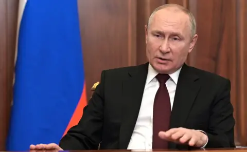 Getty Vladimir Putin pictured delivering an TV address in Feb 2022 as he announced Russia's invasion into Ukraine. He is wearing a suit and is sitting in a wood-panelled room with a Russian flag to the left of the photo.