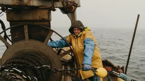 Pete Robinson A fishermen pulling a large metal chain while on a boat out at sea. His face is tense from the effort. He is wearing yellow and blue water-proof clothing with a hood over his head. The sea behind him is grey.