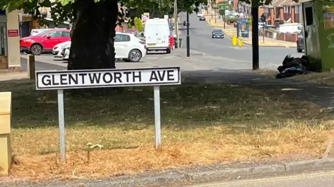 A road sign on a piece of brown grass says Glentworth Ave with a salt and grit box next to it. Behind is a large oak tree, a clothes recycling bin and a row of shops.