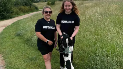 Two men in black T shirts with a logo and a black and white spaniel next to a grassy field.