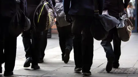 Five people are walking away from the camera and we can just see their legs and work bags. They are walking on a pavement and are waring coats and jackets.