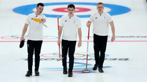 Reuters Three men in white T-shirts and black trousers and shoes walk across an Olympic curling rinkk. They are each carrying brushes and appear to be discussing the game. The ice is emblazoned with the Beijing 2022 logo.