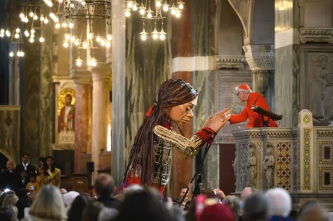 Leon Neal / Getty Images Little Amal, a giant puppet depicting a Syrian refugee girl, is addressed by the Archbishop of Westminster Cardinal Vincent Nichols as she makes her way through Westminster Cathedral in London on 25 October 2021
