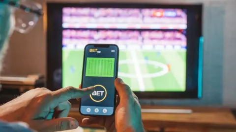 Man watching football match on TV while making a bet on a gambling app on phone