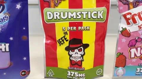 A yellow and red striped packet of sweets which mimics the Drumstick sweets packaging. There is a skeleton wearing a black hat on the front. There is a small triangle in the corner of the packet with a cannabis leaf and an 18+ circle on the opposite side.