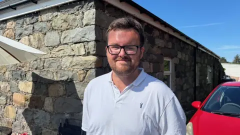 Iwan, a man with short dark hair, glasses and a beard, is looking at the camera. He is wearing a blue polo shirt and behind him is a grey stone house and a red car.