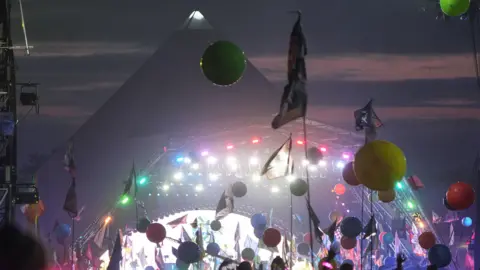 A night time shot of Glastonbury's Pyramid Stage with flags and balloons above the crowd in front of the well lit stage