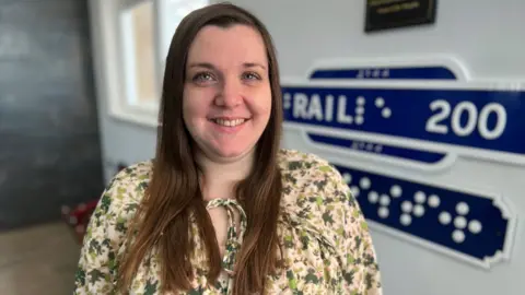 Samantha Leftwich from the Thomas Pocklington Trust. She is wearing a yellow and green floral dress, tied at the neck, and she has long brown hair. She is smiling at the camera, and is stood in front of a blue and white sign which reads Rail 200 as well as Braille dots which spell out the word Colchester